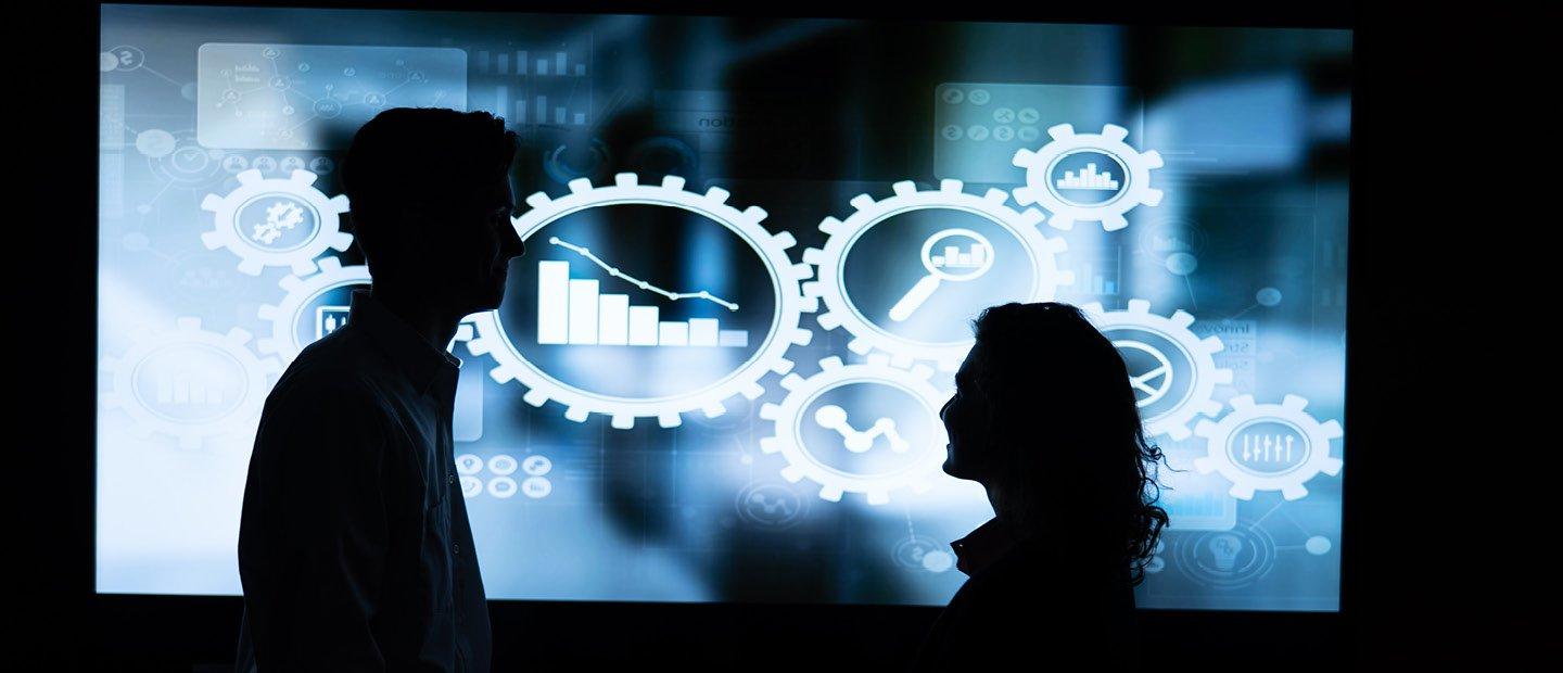 Silhouettes of two people in front of a blue screen displaying white gear and chart graphics.